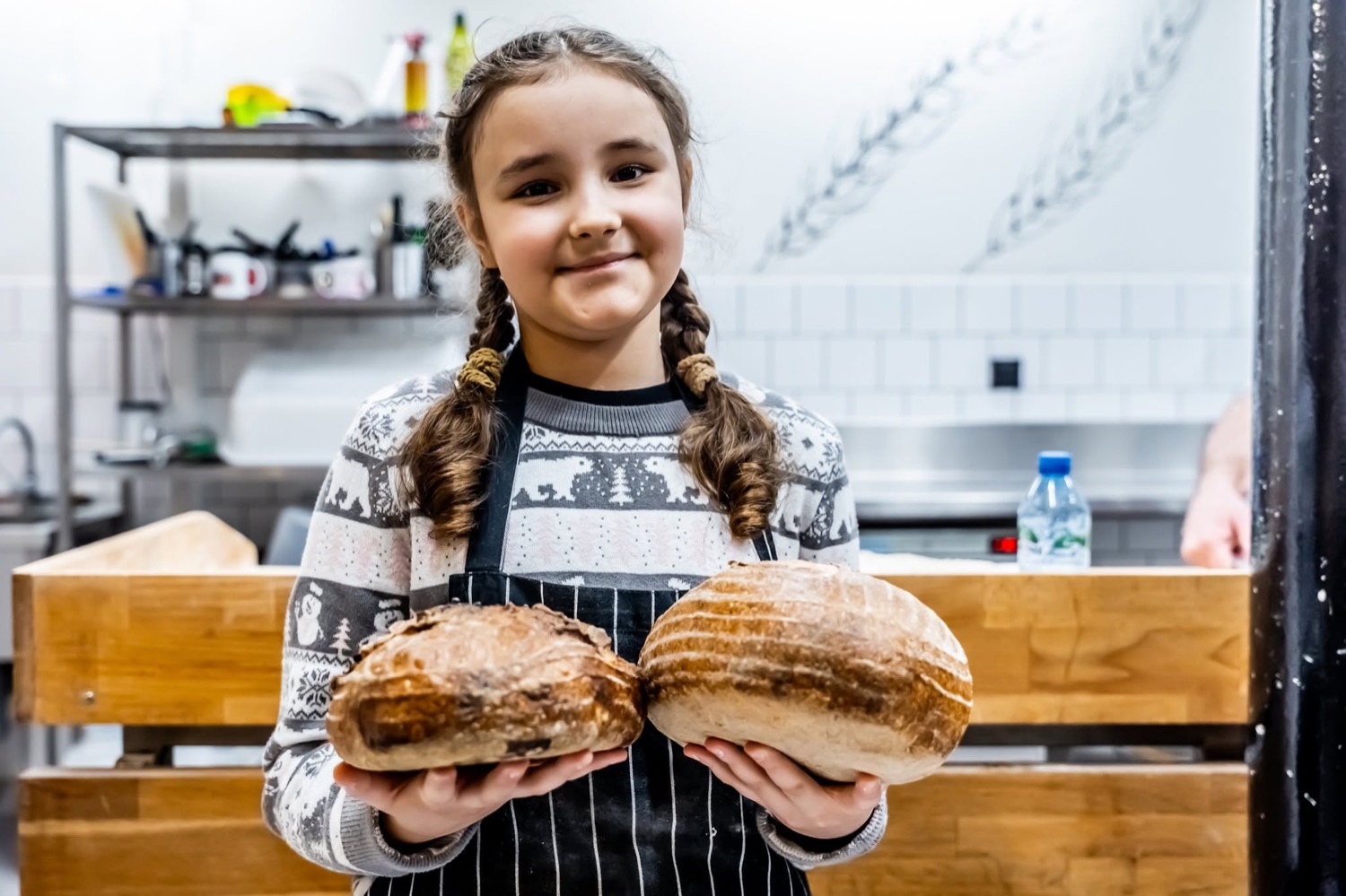 Dziewczynka w fartuchu kuchennym pokazuje dwa wypieczone chleby podczas warsztatów kulinarnych